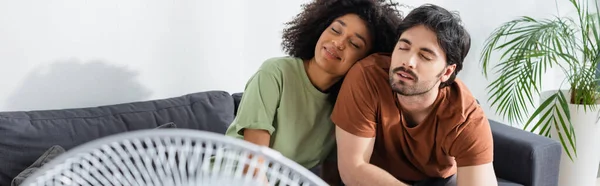 Pareja interracial complacido sentado en el sofá cerca difuminado ventilador eléctrico en la sala de estar, pancarta - foto de stock