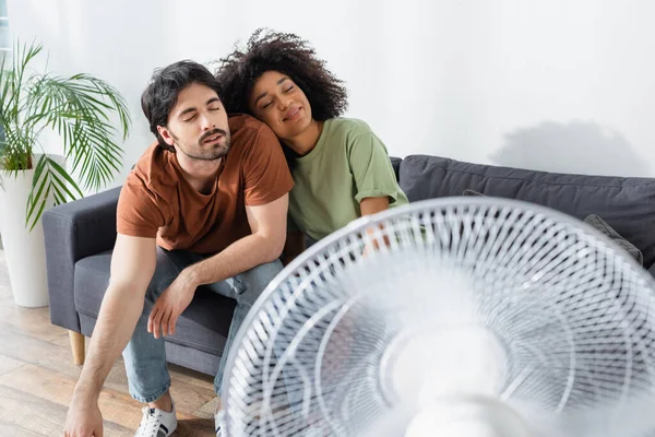 Pleased interracial couple sitting on couch near blurred electric fan in living room — Stock Photo
