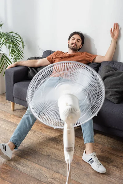 Uomo contento seduto sul divano vicino alla ventola elettrica offuscata — Foto stock