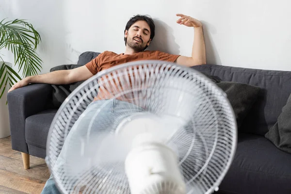 Heureux homme barbu assis sur le canapé près de ventilateur électrique flou — Photo de stock