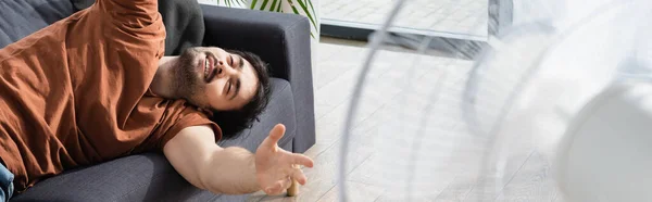 Довольный молодой человек лежит на диване возле размытого электрического вентилятора, баннер — стоковое фото