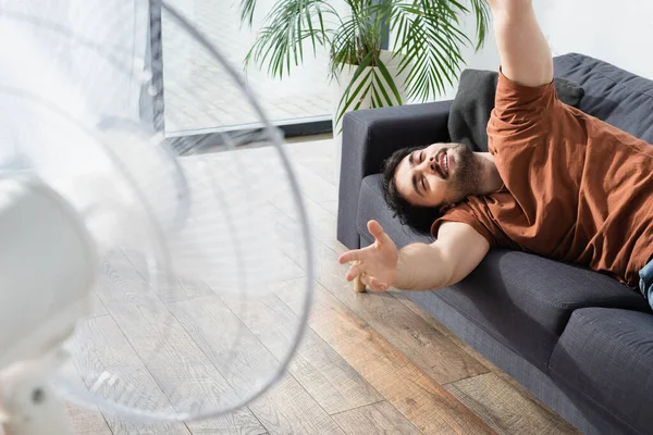 Heureux homme barbu couché sur le canapé près de ventilateur électrique flou — Photo de stock