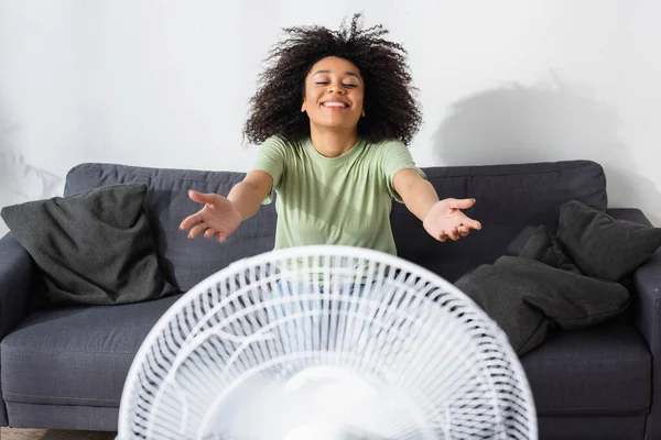 Joyeuse femme afro-américaine assise avec les mains tendues sur le canapé près d'un ventilateur électrique flou — Photo de stock