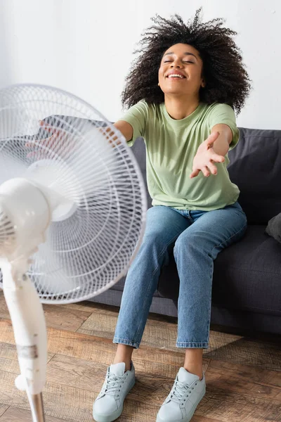 Piacere afroamericano donna seduta con le mani tese sul divano vicino sfocato ventilatore elettrico — Foto stock