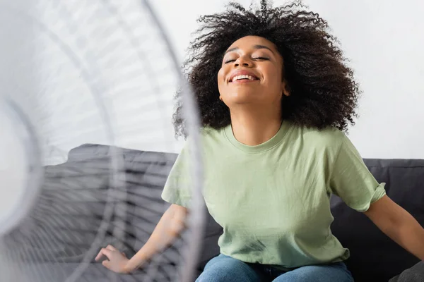 Heureux afro-américaine femme assise sur le canapé près de ventilateur électrique floue — Photo de stock