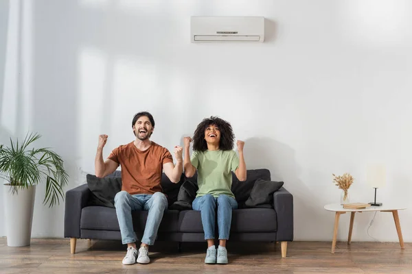 Feliz alegria multiétnica enquanto sentado no sofá na sala de estar moderna, conceito hvac — Fotografia de Stock