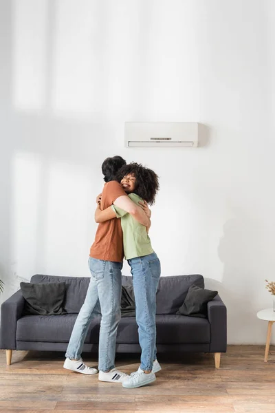 Fröhliches multiethnisches Umarmen in der Nähe einer festen Klimaanlage im modernen Wohnzimmer — Stockfoto