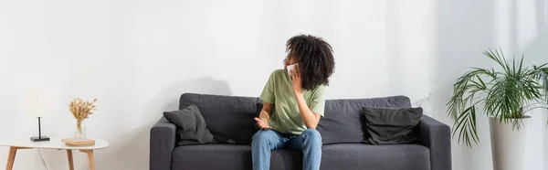Недовольная африканская американка разговаривает на смартфоне и смотрит вверх в гостиной, баннер — стоковое фото