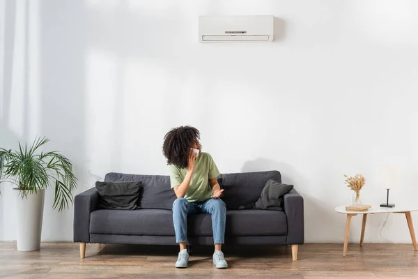 Unzufriedene Afroamerikanerin spricht auf Smartphone und blickt auf Klimaanlage im Wohnzimmer — Stockfoto