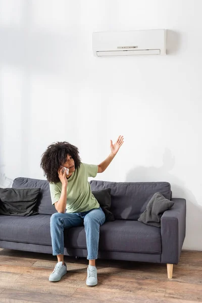Femme afro-américaine mécontente parlant sur smartphone et pointant vers climatiseur cassé dans le salon — Photo de stock
