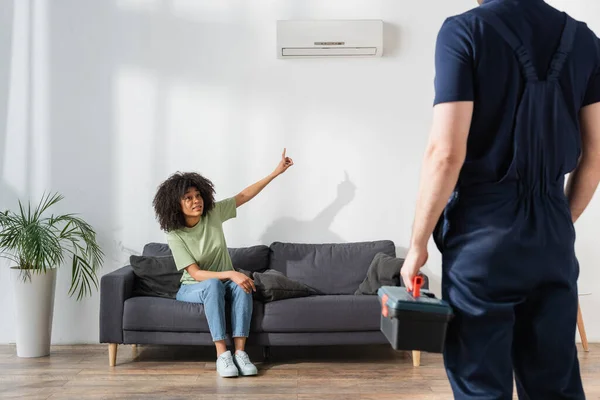 Mujer afroamericana rizada apuntando al acondicionador de aire roto cerca de handyman con caja de herramientas - foto de stock