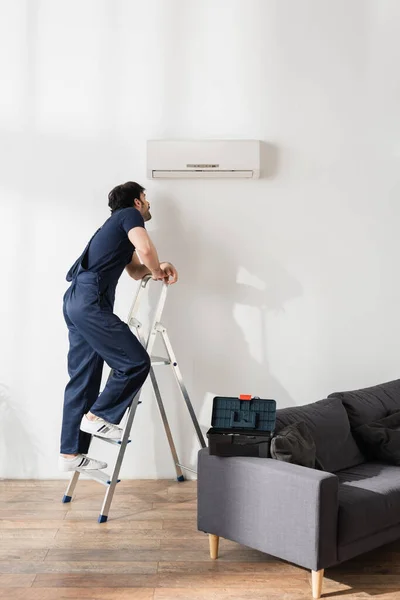 Bearded handyman in overalls standing on ladder and looking at broken air conditioner — Stock Photo