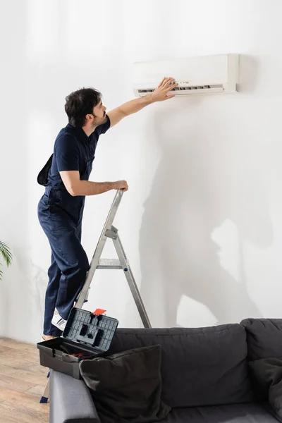 Homme à tout faire barbu en salopette debout sur l'échelle et fixer climatiseur cassé — Photo de stock