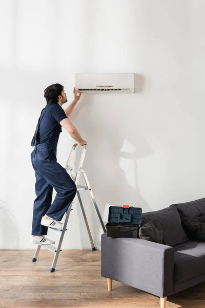 Handyman barbudo em macacão de pé na escada enquanto fixa o ar condicionado quebrado — Fotografia de Stock
