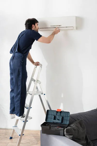 Junger Handwerker in Overalls steht auf Leiter, während er kaputte Klimaanlage repariert — Stockfoto