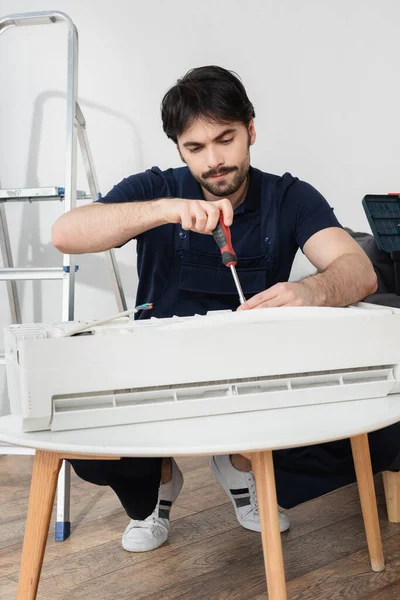 Fokussierter Handwerker in Overalls, der Schraubenzieher hält, während er kaputte Klimaanlage repariert — Stockfoto