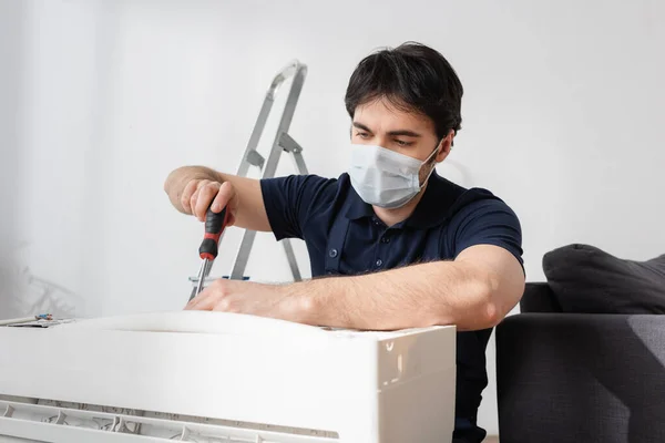 Reparateur in medizinischer Maske hält Schraubenzieher, während er defekte Klimaanlage repariert — Stockfoto