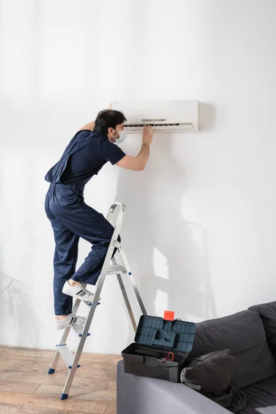 Reparateur mit medizinischer Maske steht auf Leiter und befestigt Klimaanlage im Wohnzimmer — Stockfoto