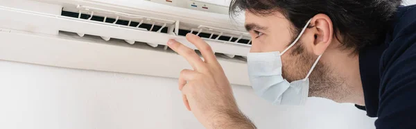 Faz-tudo em máscara médica gesticulando perto de ar condicionado enquanto verifica o controle do clima, banner — Fotografia de Stock