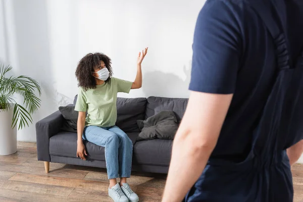 African american woman in medical mask pointing with hand near blurred handyman — Stock Photo