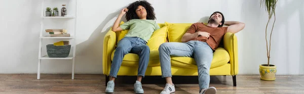 Couple multiethnique épuisé assis sur un canapé jaune et souffrant de chaleur en été, bannière — Photo de stock