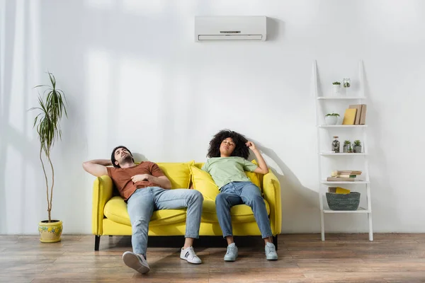 Pareja multiétnica agotada sentada en un sofá amarillo y sufriendo de calor en verano - foto de stock