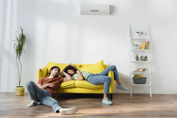 Tired multiethnic couple sitting on yellow sofa and suffering from heat in summer — Stock Photo