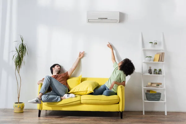 Multiethnic couple sitting on yellow sofa and reaching air conditioner while suffering from heat in summer — Stock Photo