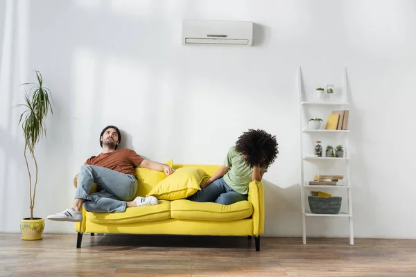Exhausted interracial couple sitting on sofa and suffering from heat in summer — Stock Photo