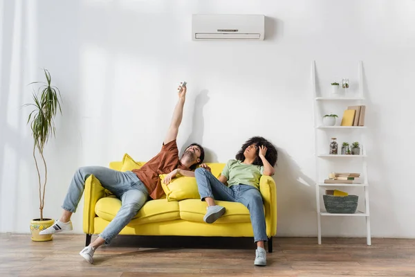 Homem segurando controlador remoto perto de ar condicionado moderno e afro-americano namorada sofrendo de calor — Fotografia de Stock