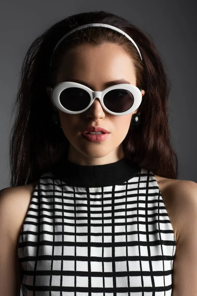 Молода жінка в передній смузі і стильні сонцезахисні окуляри дивиться на камеру ізольовані на сірому — стокове фото