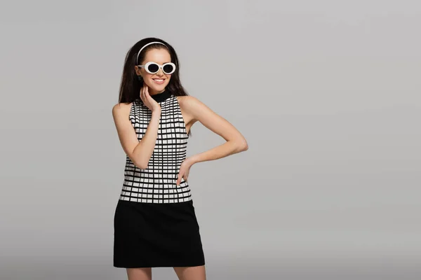 Модна молода модель в сонцезахисних окулярах, головний убір і ретро вбрання посміхаються ізольовані на сірому — стокове фото