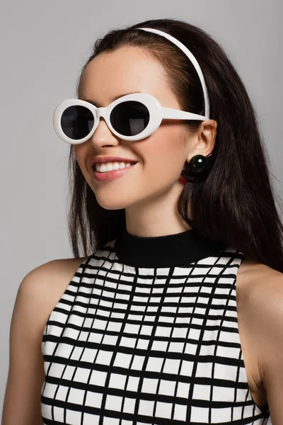 Trendy young model in sunglasses and headband smiling isolated on grey — Stock Photo