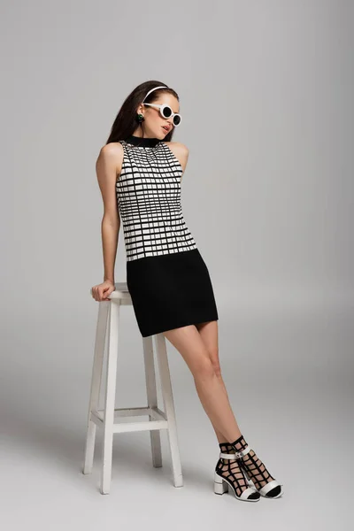 Full length of young woman in retro outfit and sunglasses leaning on high stool while posing on grey — Stock Photo