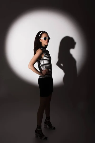 Volle Länge der Beleuchtung auf stilvolle Modell in Sonnenbrille posiert mit der Hand auf Hüfte auf grau — Stockfoto