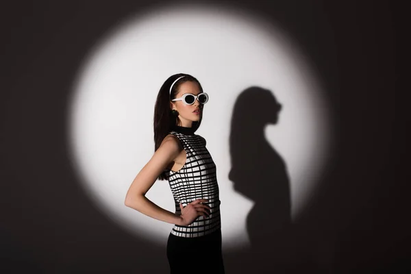 Lighting on stylish model in retro sunglasses posing with hand on hip on grey — Stock Photo