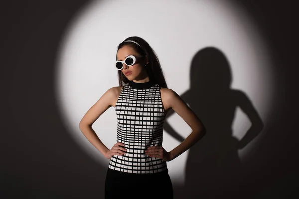 Iluminación en el modelo con estilo en gafas de sol posando con las manos en las caderas en gris - foto de stock