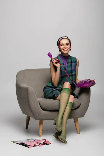 Lächelnde junge Frau mit Telefon im Sessel neben Magazin auf dem Boden auf grau — Stockfoto