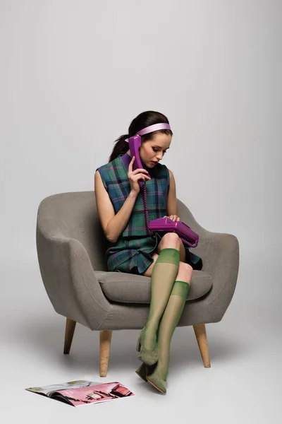 Красива молода жінка говорить на старовинному телефоні, сидячи в кріслі біля журналу на підлозі на сірому — стокове фото