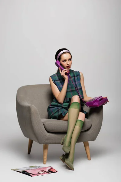 Молодая женщина разговаривает по старинному телефону, сидя в кресле рядом с журналом на полу на сером — стоковое фото