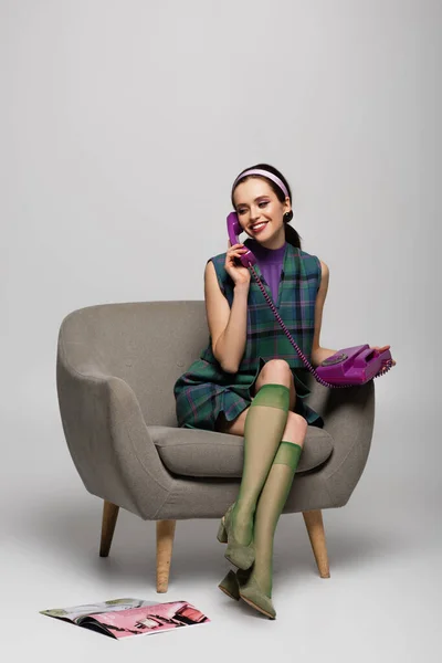 Усміхнена молода жінка говорить на ретро телефоні, сидячи в кріслі біля журналу на підлозі на сірому — стокове фото
