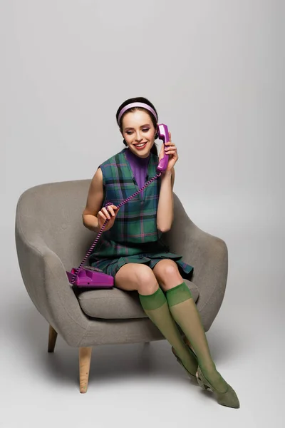 Pleased young woman speaking on phone while sitting in armchair isolated on grey — Stock Photo