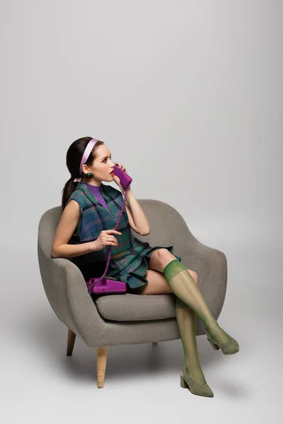 Worried young woman talking on retro telephone while sitting in armchair on grey — Stock Photo