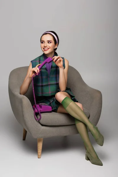 Zufriedene junge Frau mit lila Retro-Handy im Sessel auf grau — Stockfoto