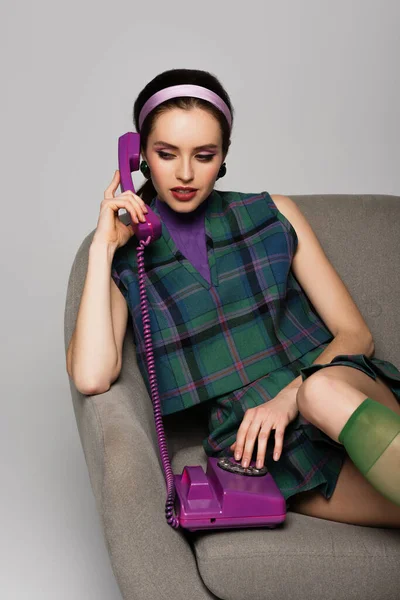 Брюнетка молода жінка говорить на фіолетовому ретро телефоні, сидячи в кріслі ізольовано на сірому — стокове фото