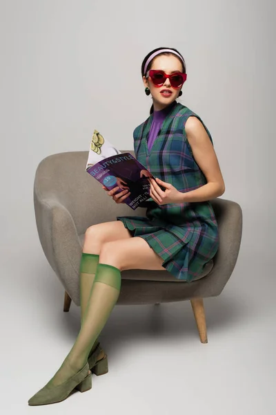 Young woman in retro sunglasses holding magazine while sitting in armchair on grey — Stock Photo