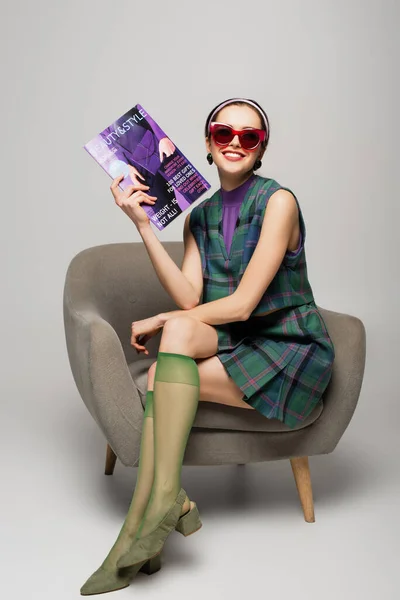 Mujer joven positiva en gafas de sol sosteniendo revista mientras está sentado en sillón en gris - foto de stock