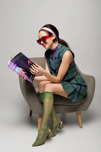 Fröhliche junge Frau mit Sonnenbrille liest Magazin, während sie im Sessel auf grau sitzt — Stockfoto