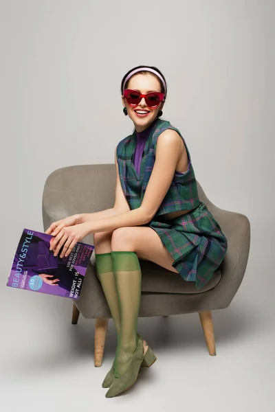 Happy young woman in sunglasses holding magazine while sitting in armchair on grey — Stock Photo