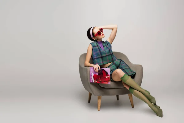 Молодая женщина в солнцезащитных очках держит журнал, позируя и сидя кресло на сером — стоковое фото